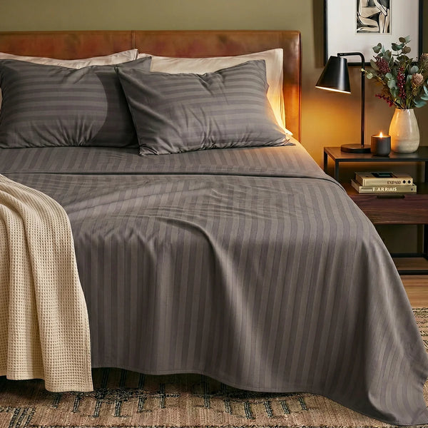 A neatly made bed with the Super Sleeper Pro Imperial Dream Sheets™ 4pc Bedding Set and two pillows sits by a wooden nightstand with a lamp, vase of flowers, candle, and books. A cream knitted blanket lies at the foot of the bed.