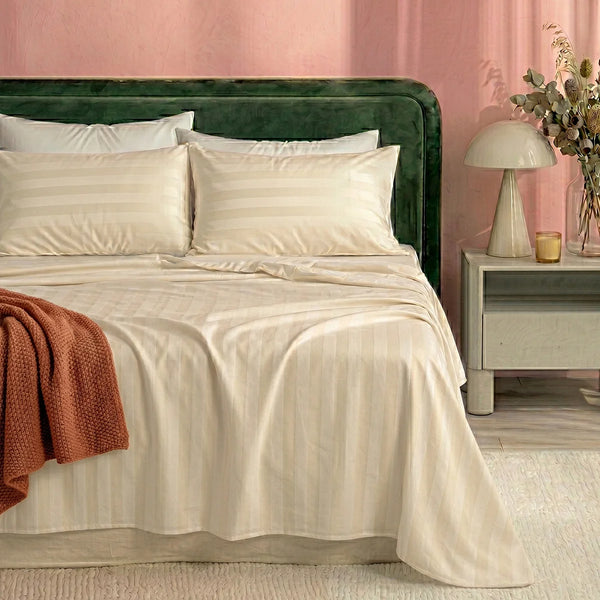 A neatly made bed with a green upholstered headboard showcases the Super Sleeper Pro Imperial Dream Sheets™ 4pc Bedding Set in cream stripes and pillows. A rust blanket is draped on one side; beside it, a nightstand holds a lamp and a vase of flowers.
