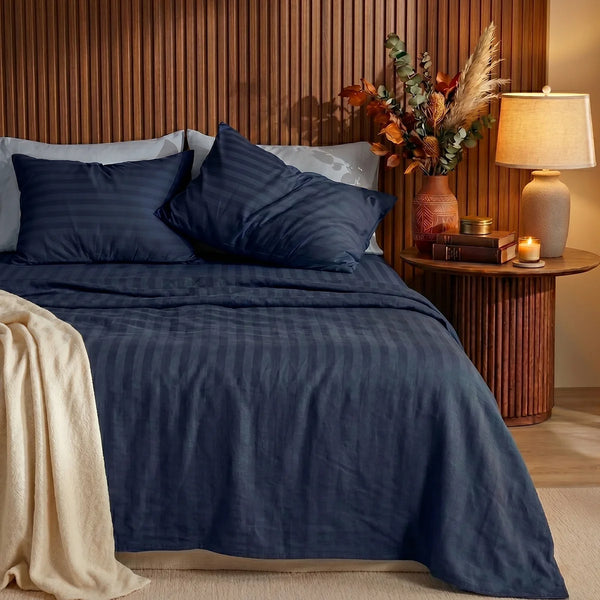 A neatly made bed showcases the Super Sleeper Pro Imperial Dream Sheets™ 4pc Bedding Set in dark blue stripes, with two pillows and a beige blanket. Against a wooden headboard, a side table holds a lamp, books, candle, and autumn foliage.