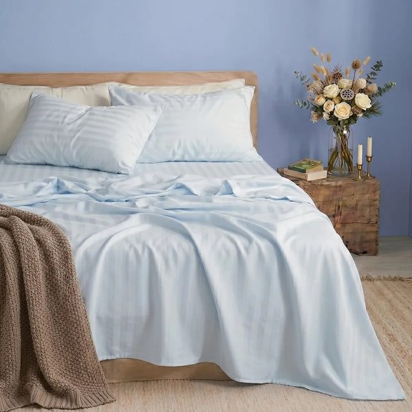 A neatly made bed with the Super Sleeper Pro Imperial Dream Sheets™ 4pc Bedding Set in light blue and two pillows sits against a light blue wall. A nightstand with flowers, candles, and books is beside the bed; a brown knitted blanket is draped over the corner.