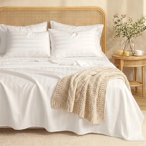 A made bed with Super Sleeper Pro's Imperial Dream Sheets™ 4pc Bedding Set in white stripes, three pillows, a beige knitted blanket, wooden headboard, and a round side table with vase, two cups, and teapot on a light rug.