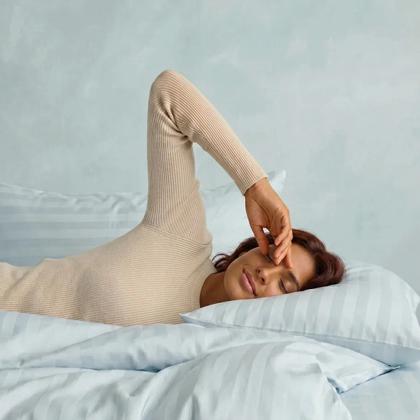 A person with long hair lies on a bed dressed in Super Sleeper Pro's Imperial Dream Sheets™ 4pc Bedding Set, featuring light blue stripes and 1200TC cotton sateen, wearing a beige ribbed long-sleeve top and resting an arm across their forehead.