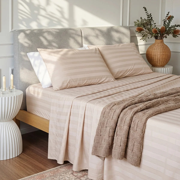 A neatly made bed with Super Sleeper Pro’s Imperial Dream Sheets™ 4pc Bedding Set in beige stripes, two pillows, a brown knit blanket, gray upholstered headboard, round white nightstand with candles, and a wicker vase of dried flowers.