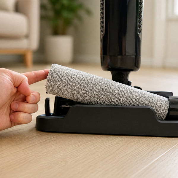 A hand removes the cylindrical roller brush from the base of the black Kleva Range FX5 Floor Washer on light wood flooring, with a blurred plant and beige furniture visible in the background.