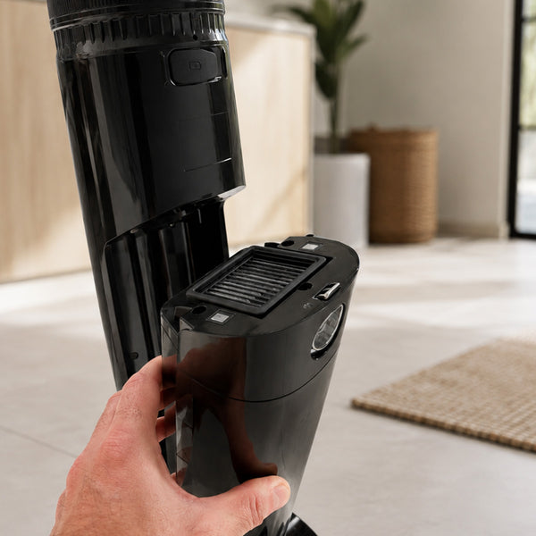 A hand is removing a rectangular, black FX5 Floor Washer - Filter Kit (2 pack) by Kleva Range from the main body of a black cordless floor washer in a modern room with a beige rug and a woven basket in the background.