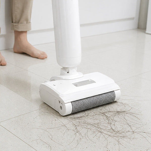 A person uses the Kleva Range FX5 Floor Washer - Roller Brush (White) to pick up clumps of hair from a light-colored tiled floor, with only their bare feet and lower legs visible in the background.