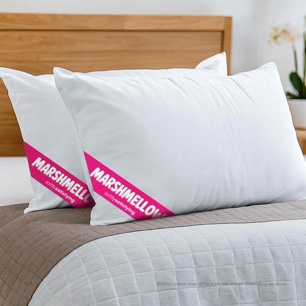 Two white Super Sleeper Pro pillows with pink "MARSHMELLOW" bands rest on a neatly made bed with a beige quilt, wooden headboard, and a Marshmellow™ Plush Mattress Topper for added comfort. Includes 2 free pillows.