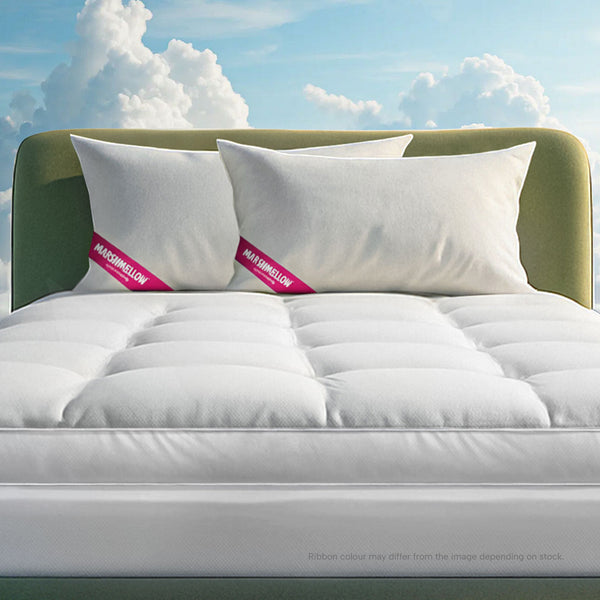 A bed featuring the Super Sleeper Pro Marshmellow™ Plush Mattress Topper and 2 free pillows, both with red and white tags, stands out with a green upholstered headboard against a blue sky and cloud backdrop.