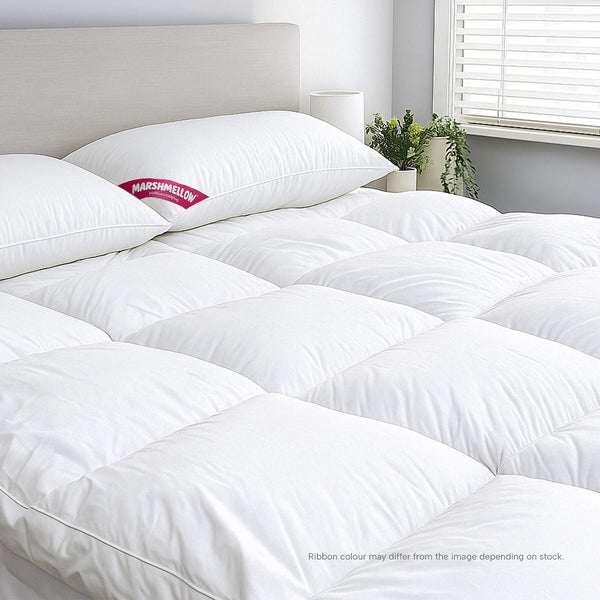 A bright bedroom features a neatly made bed with the Super Sleeper Pro Marshmellow™ Plush Mattress Topper and 2 free pillows. A plant and clock rest on the bedside table near white blinds, enhancing comfort and style.