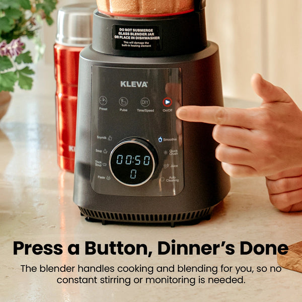 A hand presses the On/Off button on a black Kleva Range Mealio® Hot & Cold Blender with a digital display reading 00:50. The appliance sits on a kitchen counter beside a red container, showcasing its all-in-one convenience.