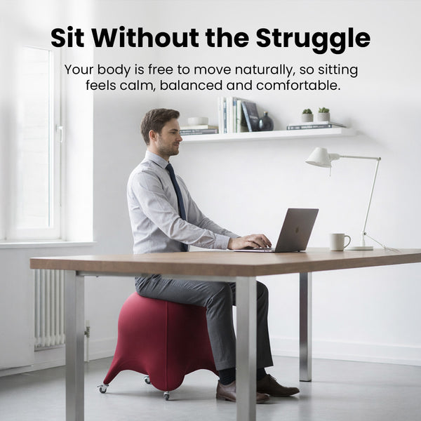 A man in business attire sits at a desk using a laptop on the KLEVA® Posture Chair by Kleva Range, designed for active sitting to improve posture and core strength. The minimalistic room has a white wall, shelf with books and decor, and a lamp.