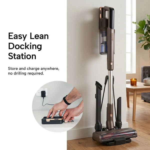 The KRAPOF® S3 Power Vac Cordless Stick Vacuum by Kleva Range stands upright on its Easy Lean Docking Station next to a wooden table; hands plug the station into a wall outlet. Store and charge anywhere—no drilling required. Includes free extra battery.