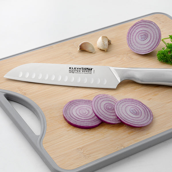 The Kleva Range KlevaCut® Master Series Professional Santoku Knife - 18cm rests on a wooden cutting board with slices of red onion, two garlic cloves, and a sprig of parsley.