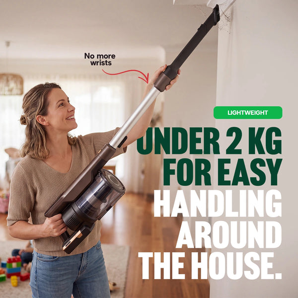A woman uses the KRAPOF® Power Vac S3 Cordless Stick Vacuum by Kleva Range to clean a corner of the ceiling in a living room. Text on image highlights “Under 2 kg for easy handling around the house” and “No more wrist strain.”.