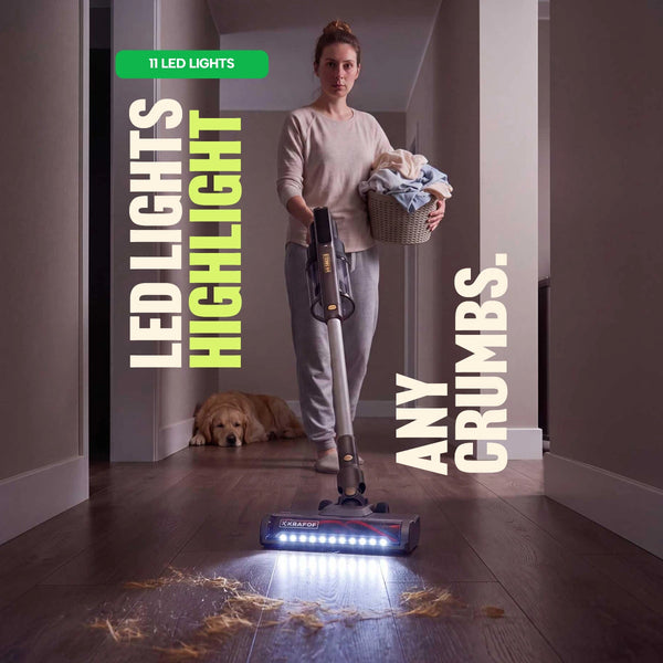 A person carries a laundry basket and uses the Kleva Range KRAPOF® Power Vac S3 Cordless Stick Vacuum + Extra FREE Second Battery to clean crumbs from a hardwood floor, while a dog lies in the background. Text reads: 11 LED lights highlight every crumb.