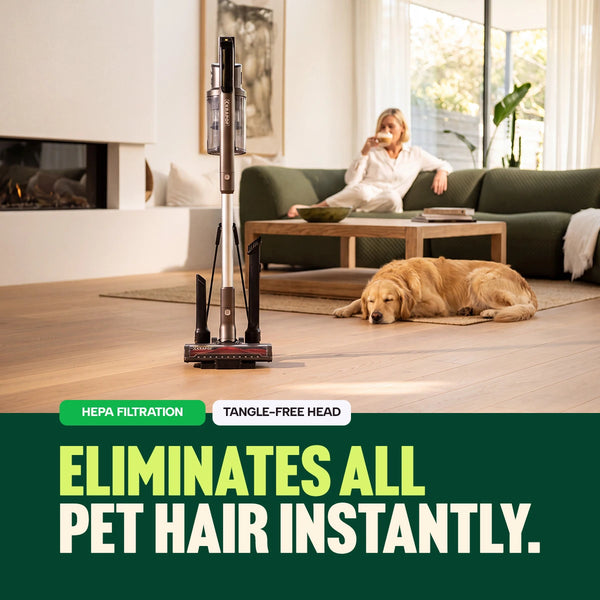 A KRAPOF® Power Vac S3 Cordless Stick Vacuum from Kleva Range stands on a wooden floor in a living room. A woman relaxes on a green couch while her golden retriever lies nearby. Text reads: Instantly eliminates all pet hair. Advanced HEPA filtration. Tangle-free head for effortless pet hair removal. Includes an extra FREE second battery!.