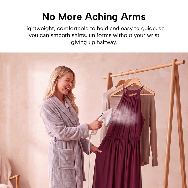 A woman in a robe uses the KLEVA® Crease Boss Steam Iron by Kleva Range as an effective wrinkle remover to smooth out a sleeveless burgundy dress hanging on a wooden rack, with a beige sweater draped over the rack.