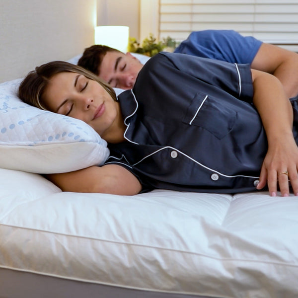 A woman and a man are asleep in bed, with the woman in the foreground resting on a Super Sleeper Pro Marshmellow™ Plush Mattress Topper and pillow. A lamp and greenery are visible in the background.