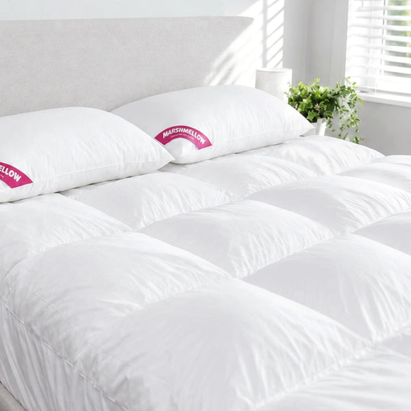 A neatly made bed features the Super Sleeper Pro Marshmellow™ Plush Mattress Topper and two free pillows. Sunlight filters through window blinds, while a green potted plant on a white nightstand enhances the cozy bedroom ambiance.