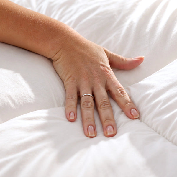 A hand with a simple ring rests on the Marshmellow™ Plush Mattress Topper by Super Sleeper Pro, evoking comfort. This set includes 2 free pillows for a cozy bed experience.