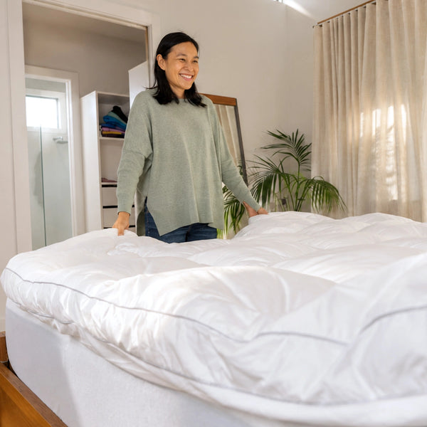 A woman smiles as she smooths out the Marshmellow™ Plush Mattress Topper by Super Sleeper Pro on her bed. Sunlight streams into the cozy room, which features beige curtains and a neat closet. Two free pillows complete her comfy sleep setup.