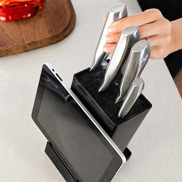 A person inserts a Kleva Range KlevaCut® Master Series TV Special 4pc Knife Set stainless steel knife into the sleek black universal knife block, which also features a front slot for holding a tablet. On the countertop in the background, a cutting board with sliced red peppers is visible.