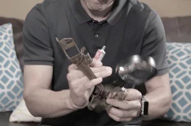 A person in a dark polo shirt applies lubricant to a large adjustable wrench while holding a magnifying glass. Two patterned pillows and a couch are visible in the background.