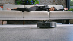 A woman stretched out on the couch while the robot vacuums the floor in front of her.
